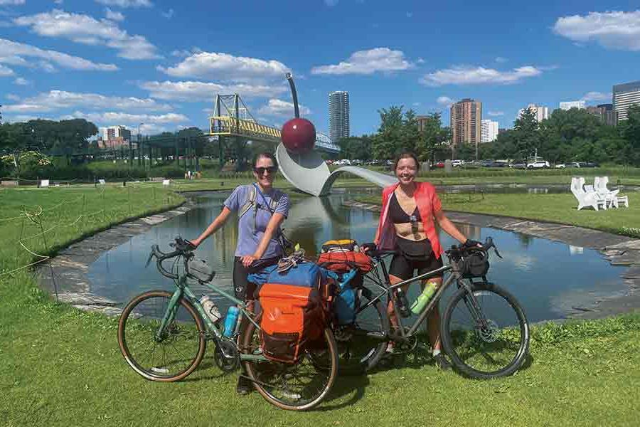 Brenna and her biking companion pose with their bikes in front of a pond with blue sky and fluffy clouds behind them